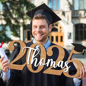 May include: A person wearing a black graduation gown and cap holds a wooden sign with the year "2023" and the name "Thomas" in white lettering. The sign also features a black graduation cap icon.