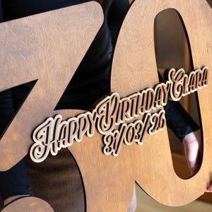 Op de afbeelding: Een grote houten 70e verjaardagsdecoratie. Het nummer is lichtbruin met een uitgesneden ontwerp. De woorden "HAPPY BIRTHDAY Clara!" en de datum 21/03/26 zijn gelaagd.