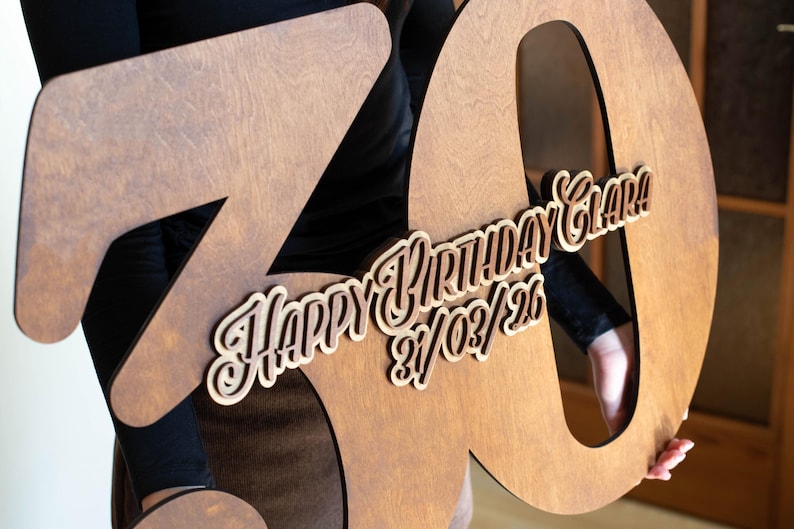 Op de afbeelding: Een grote houten 70e verjaardagsdecoratie. Het nummer is lichtbruin met een uitgesneden ontwerp. De woorden "HAPPY BIRTHDAY Clara!" en de datum 21/03/26 zijn gelaagd.