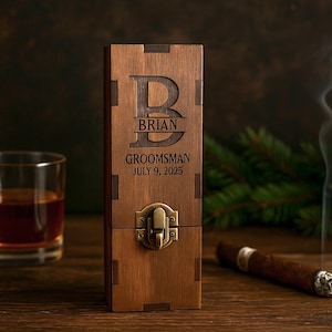 May include: A wooden box with the name "Brian", the word "Groomsman", and the date "July 9, 2025" engraved on it. A glass of amber liquid, a lit cigar, and smoke are also visible. The box has a metal clasp.