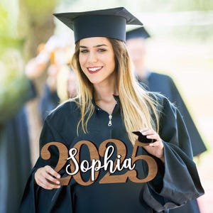 May include: A wooden graduation sign with the year 2025 and the name Sophia written in white script. The sign is held by a person wearing a black graduation gown and cap.