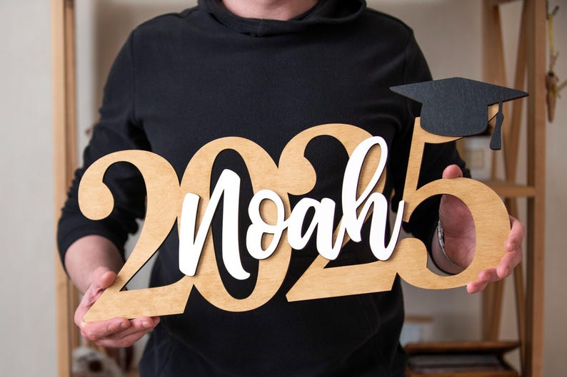 May include: A wooden sign with the year 2025 and the name Noah in white lettering. A black graduation cap is on top of the 5. The sign is held by a person.