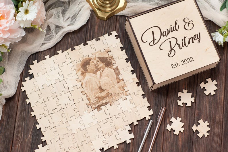 Peut inclure: Une bo&icirc;te &agrave; puzzle en bois avec une inscription personnalis&eacute;e "David & Britney Est. 2022" et une photo de couple. La bo&icirc;te &agrave; puzzle est sur une surface en bois avec une pi&egrave;ce de puzzle blanche et un stylo.