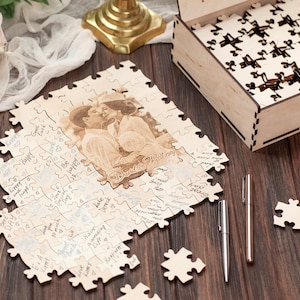 Peut inclure: Un puzzle en bois avec une photo de couple imprim&eacute;e dessus. Le puzzle est assembl&eacute; et a une bo&icirc;te en bois &agrave; c&ocirc;t&eacute;. Les pi&egrave;ces du puzzle sont dispers&eacute;es autour de la bo&icirc;te. Deux stylos sont &eacute;galement sur la table.