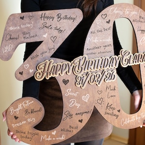 Op de afbeelding: Een houten "30" verjaardagsbord met de woorden "Happy Birthday Clara" en de datum "21/03/26" in een decoratief lettertype. Het bord is versierd met harten en verjaardagswensen. Een persoon houdt het bord vast.