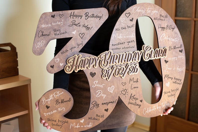 Op de afbeelding: Een houten "30" verjaardagsbord met de woorden "Happy Birthday Clara" en de datum "21/03/26" in een decoratief lettertype. Het bord is versierd met harten en verjaardagswensen. Een persoon houdt het bord vast.