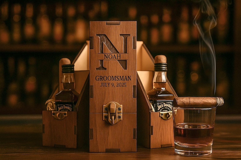 May include: A wooden gift box with the name "NOAH" and the text "Groomsman July 9, 2025" engraved on the front. The box holds two miniature whiskey bottles and a glass of whiskey with a cigar resting on the rim.