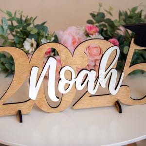 May include: Wooden sign with the year 2025 and the name Noah in white lettering. A black graduation cap sits on top of the 5.