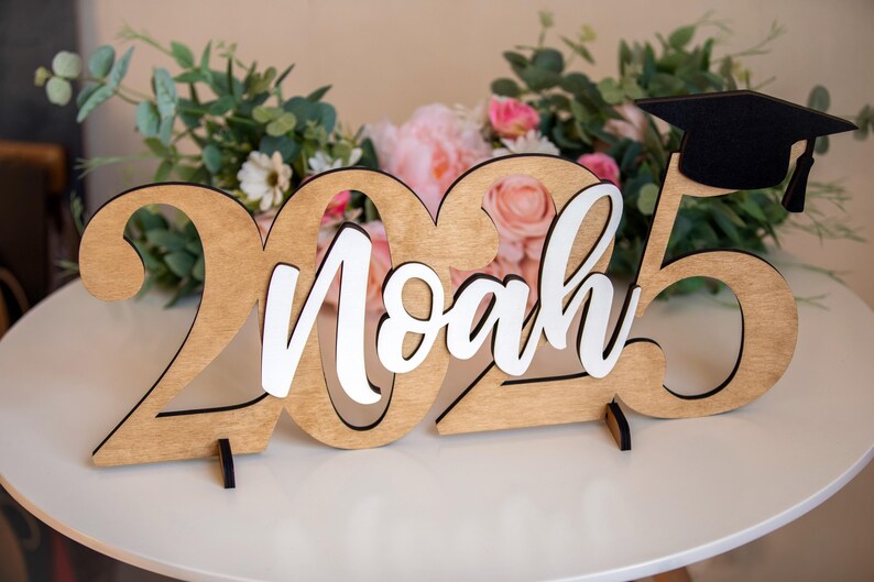 May include: Wooden sign with the year 2025 and the name Noah in white lettering. A black graduation cap sits on top of the 5.