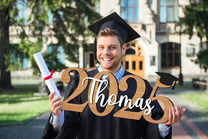 May include: A person wearing a black graduation gown and cap holds a wooden sign with the year "2023" and the name "Thomas" in white lettering. The sign also features a black graduation cap icon.