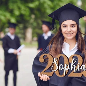 May include: A woman in a black graduation gown holds a wooden sign with the year 2025 and the name Sophia written in white. The sign is decorated with a small graduation cap.
