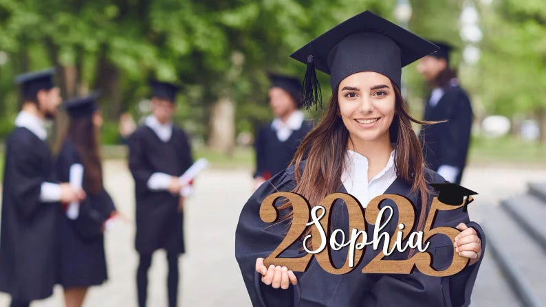 May include: A woman in a black graduation gown holds a wooden sign with the year 2025 and the name Sophia written in white. The sign is decorated with a small graduation cap.