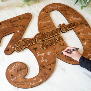 Op de afbeelding: Houten 40e verjaardagsbord met de woorden "Happy Birthday" en "Clara" in een decoratief lettertype. Het bord is bruin met witte tekst en hartjes, en bevat de datum 21/03/26. Een hand schrijft met een zwarte pen op het bord.