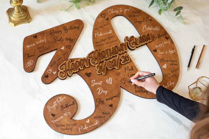 Op de afbeelding: Houten 40e verjaardagsbord met de woorden "Happy Birthday" en "Clara" in een decoratief lettertype. Het bord is bruin met witte tekst en hartjes, en bevat de datum 21/03/26. Een hand schrijft met een zwarte pen op het bord.