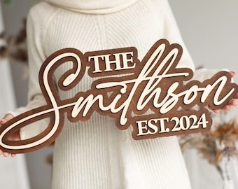 May include: A brown wooden sign with the text "The Smithson EST. 2024" in white lettering. The sign is held by a person wearing a white sweater.