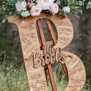 Pu&ograve; includere: Un cartello di legno a forma delle lettere "B" ed "R" con il testo "The Brooks" al centro. Il cartello &egrave; decorato con fiori e ha messaggi scritti a mano.