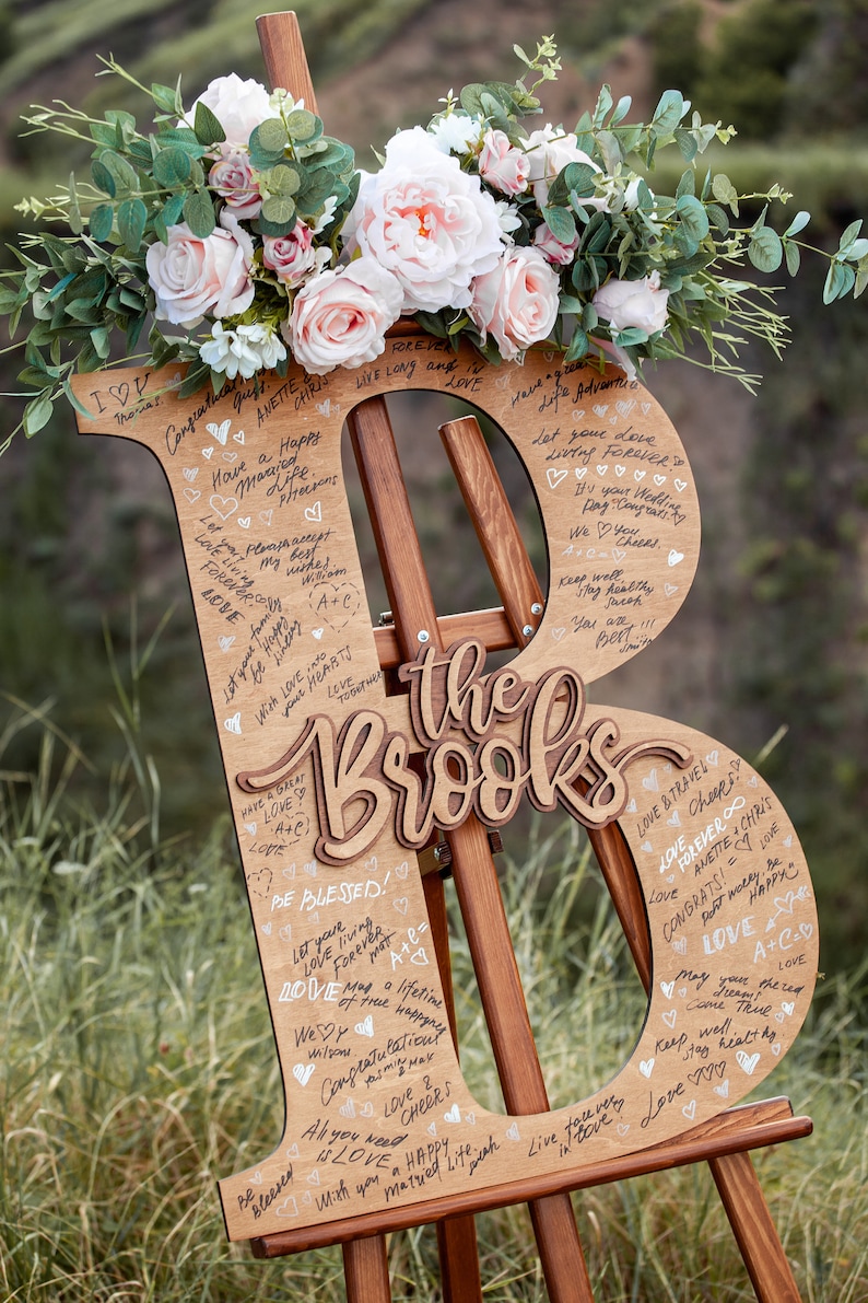 Pu&ograve; includere: Un cartello di legno a forma delle lettere "B" ed "R" con il testo "The Brooks" al centro. Il cartello &egrave; decorato con fiori e ha messaggi scritti a mano.
