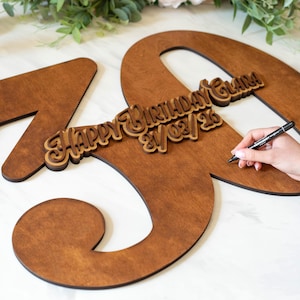 Op de afbeelding: Houten 70e verjaardagsbord in een warme bruine tint. Het bord bevat een kleinere houten inscriptie met de tekst "Happy Birthday Clara 21/03/26". Een hand houdt een pen vast, vermoedelijk om het bord te ondertekenen.