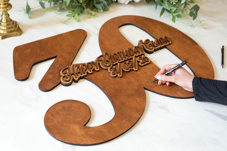 Op de afbeelding: Houten 70e verjaardagsbord in een warme bruine tint. Het bord bevat een kleinere houten inscriptie met de tekst "Happy Birthday Clara 21/03/26". Een hand houdt een pen vast, vermoedelijk om het bord te ondertekenen.