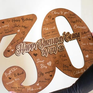 Op de afbeelding: Een houten verjaardagsdecoratie in de vorm van het cijfer 30. Het bruine cijfer heeft lichtere houten accenten en de woorden "Happy Birthday Clara" en de datum 31/03/26. Het is versierd met hartjes en verjaardagswensen.