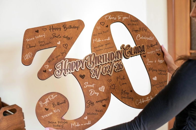 Op de afbeelding: Een houten verjaardagsdecoratie in de vorm van het cijfer 30. Het bruine cijfer heeft lichtere houten accenten en de woorden "Happy Birthday Clara" en de datum 31/03/26. Het is versierd met hartjes en verjaardagswensen.