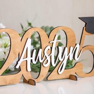 May include: Wooden graduation sign with the year 2025 and the name Austyn in white lettering. A black graduation cap is on top of the sign.