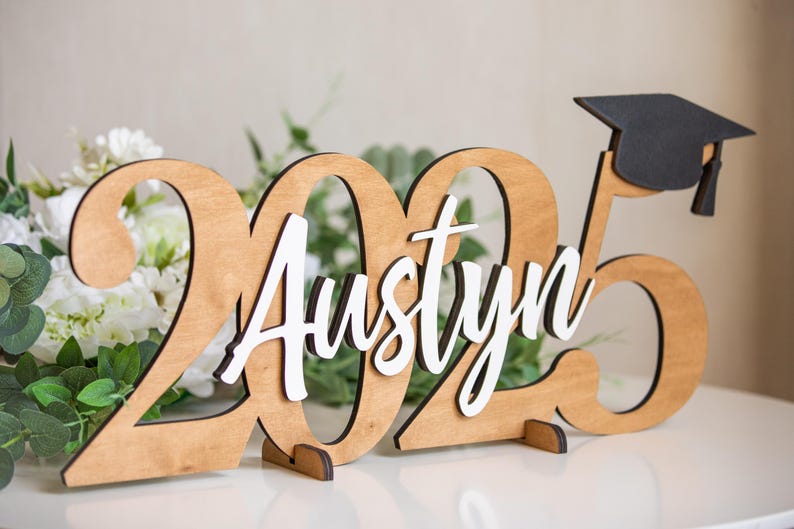 May include: Wooden graduation sign with the year 2025 and the name Austyn in white lettering. A black graduation cap is on top of the sign.