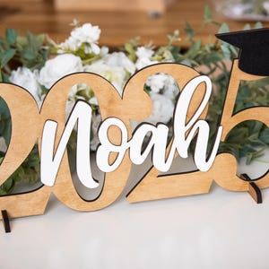 May include: Wooden 2023 graduation sign with the name Noah in white lettering and a black graduation cap on top.