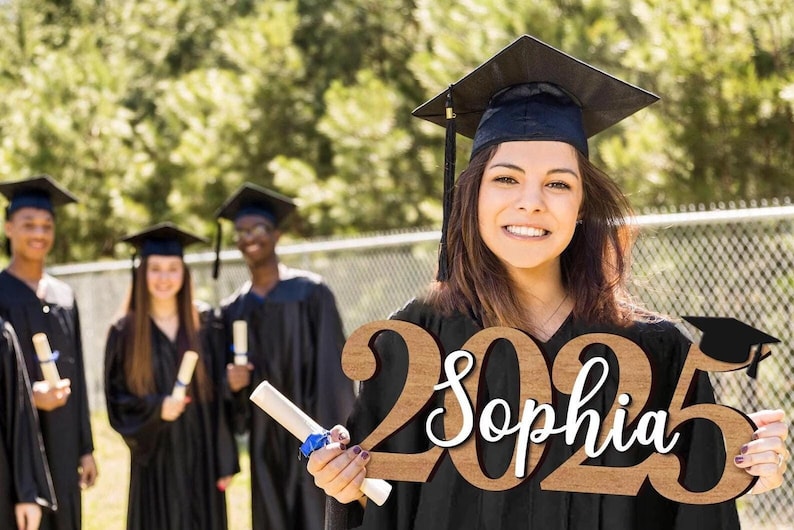 May include: A brown wooden sign with the year 2025 and the name Sophia written in white cursive. The sign is being held by a person wearing a black graduation gown and cap.