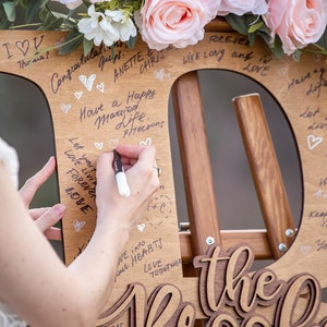 Pu&ograve; includere: Un cartello di legno a forma della lettera "D" con le parole "the Brook" scritte in corsivo. Il cartello &egrave; decorato con fiori rosa e ha diversi messaggi scritti su di esso con un pennarello nero.