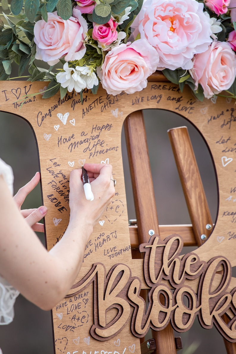 Pu&ograve; includere: Un cartello di legno a forma della lettera "D" con le parole "the Brook" scritte in corsivo. Il cartello &egrave; decorato con fiori rosa e ha diversi messaggi scritti su di esso con un pennarello nero.