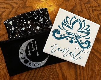 May include: Two fabric pouches with different designs. One pouch is black with a white moon and stars pattern. The other pouch is black with a white crescent moon and hanging ornaments. A white ceramic tile with a blue lotus flower design and the word "Namaste" is on top of the pouches.