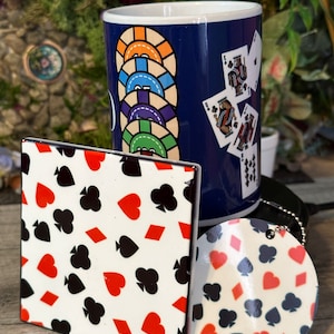 May include: A blue ceramic mug with a design of playing cards and poker chips. The mug is sitting on a white coaster with a black and red playing card pattern. There is a round coaster with the same pattern next to the mug.