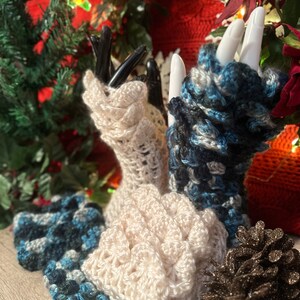 May include: A set of three crocheted fingerless gloves in white, blue, and black. The gloves are displayed on a wooden surface with a pine cone.