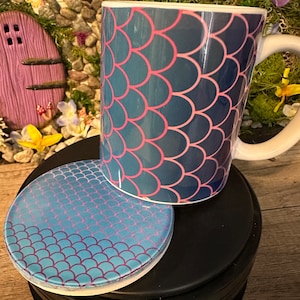 May include: A blue and pink ceramic mug with a mermaid scale pattern. The mug is sitting on a matching coaster.