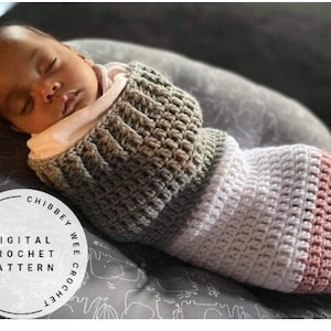 Peut inclure: Un bébé emmailloté dans une couverture en crochet gris, blanc et rose. La couverture est réalisée avec un point texturé et présente un motif rayé. Le bébé dort paisiblement. L'image comprend le texte "Digital Crochet Pattern" dans un cercle.