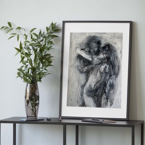 May include: Abstract black and white artwork in a black frame. The painting depicts two figures embracing, with broad brushstrokes creating a sense of movement. The artwork is displayed on a dark gray table, next to a vase with green leaves.