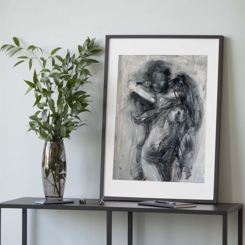 May include: Abstract black and white artwork in a black frame. The painting depicts two figures embracing, with broad brushstrokes creating a sense of movement. The artwork is displayed on a dark gray table, next to a vase with green leaves.