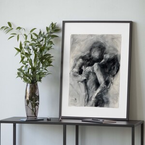 May include: Black and white abstract art print of two figures embracing. The artwork is framed in a black frame and displayed on a dark gray table. A vase with green leaves is to the left of the artwork.