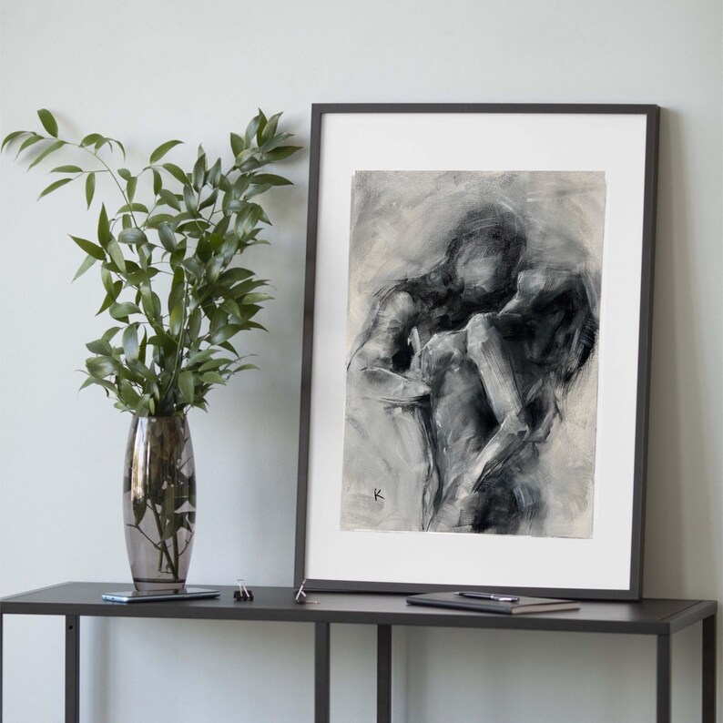 May include: Black and white abstract art print of two figures embracing. The artwork is framed in a black frame and displayed on a dark gray table. A vase with green leaves is to the left of the artwork.