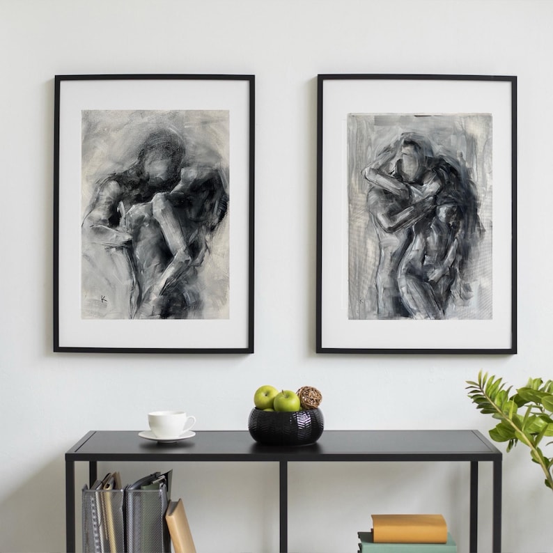 May include: Two framed black and white abstract art prints depicting figures in intimate embraces. The artwork is displayed on a white wall above a black table with a decorative bowl and a cup and saucer.