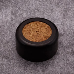 Puede incluir: Un tapón de botella redondo negro con un tapón de corcho.