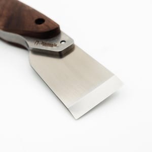 May include: A close-up of a small, stainless steel scraper with a wooden handle. The scraper has a beveled edge and is designed for scraping and cleaning.