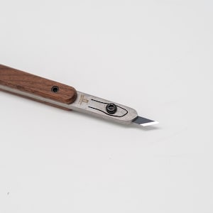 May include: A close-up of a craft knife with a wooden handle and a silver blade. The blade is angled and sharp, with a black screw securing it. The handle is a dark brown color.