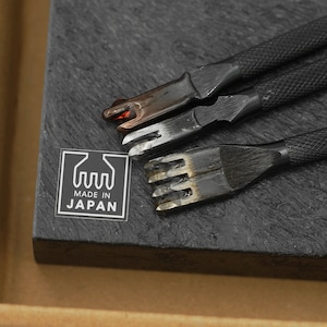 May include: Three black leather crafting tools with metal tips on a black slate surface. The tools are arranged in a row, with the tips facing the viewer. The slate surface has a label that reads "MADE IN JAPAN".