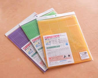 May include: Three self-healing cutting mats in purple, green, and yellow. The mats are labeled "NT Cutting Mat" and have grid lines. The mats are available in sizes A5 (22 x 15 cm), A4 (30 x 22 cm), and A3 (45 x 30 cm).