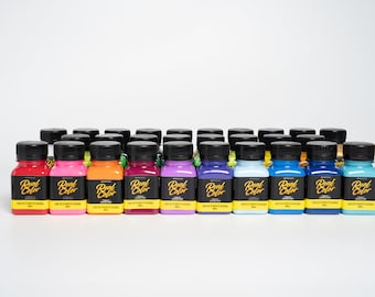 Könnte beinhalten: Eine Sammlung von 24 kleinen Farbflaschen, jede mit einem schwarzen Deckel und einem gelben Etikett mit der Aufschrift "Real Color". Die Flaschen sind in Reihen zu sechs angeordnet, wobei jede Reihe eine andere Farbe von Farbe zeigt.