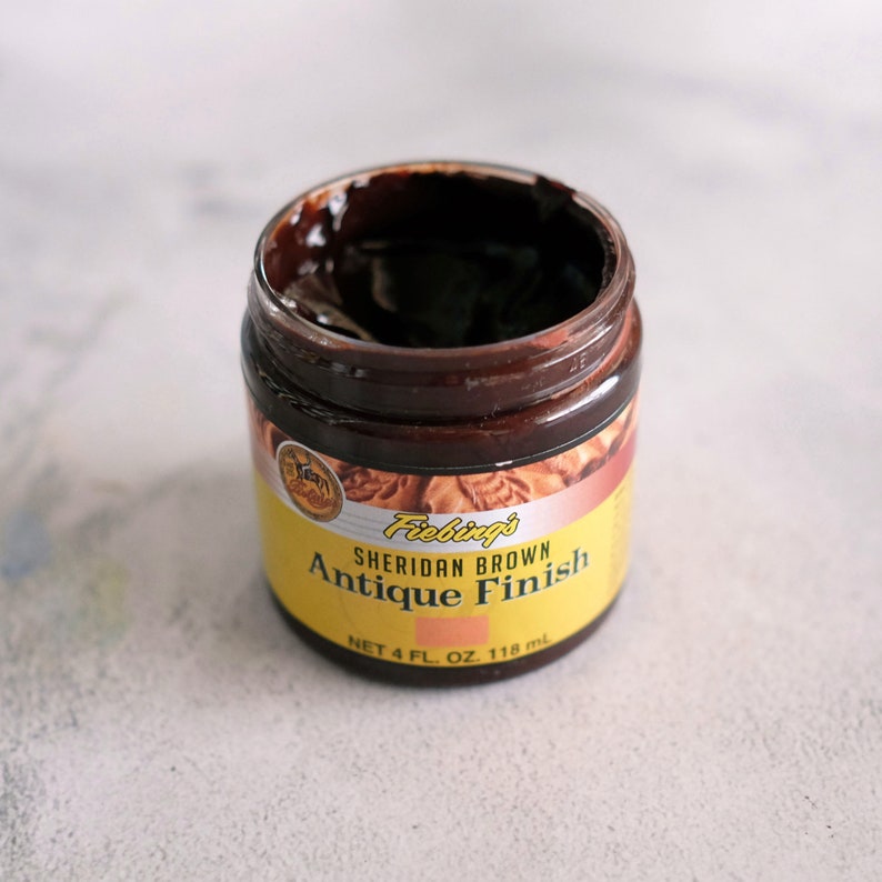 May include: A glass jar of Fiebing's Sheridan Brown Antique Finish leather dye. The jar is yellow with black text and a brown lid. The dye is a dark brown color.