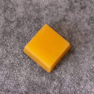 Puede incluir: Una barra de cera de abejas de color amarillo brillante con forma cuadrada.