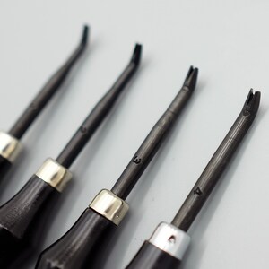 May include: Four black leather working tools with silver metal tips and numbered handles. The tools are arranged in a row on a white surface.